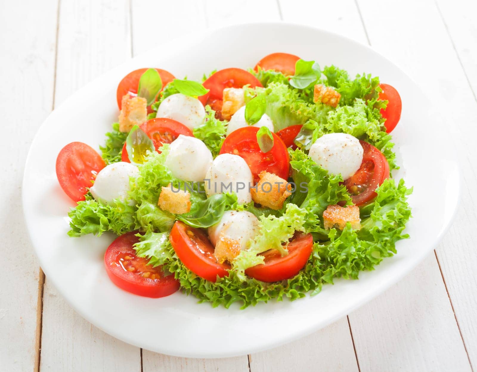 Fresh Italian salad with mozzarella cheese pearls on a bed of frilly lettuce with tomatoes and friend crunchy bread croutons served on a plate on white boards