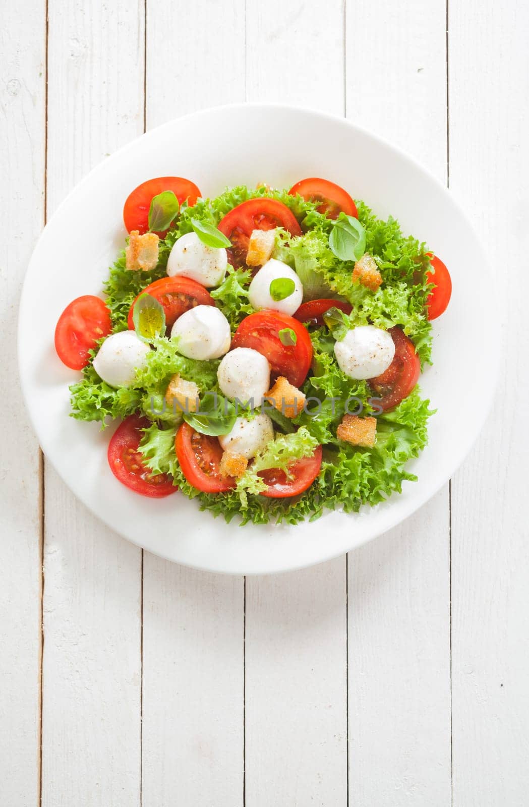 Italian Caprese salad with mozzarella pearls and tomato on a bed of lettuce garnished with golden crunchy friend bread croutons, overhead view on white boards with copyspace