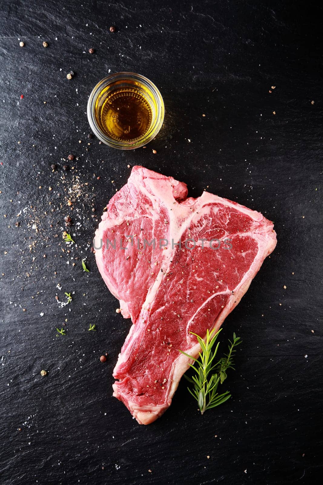 Preparing a t-bone steak for grilling with an overhead view of a lean steak seasoned with spice and herbs with a glass of bourbon or whiskey alongside