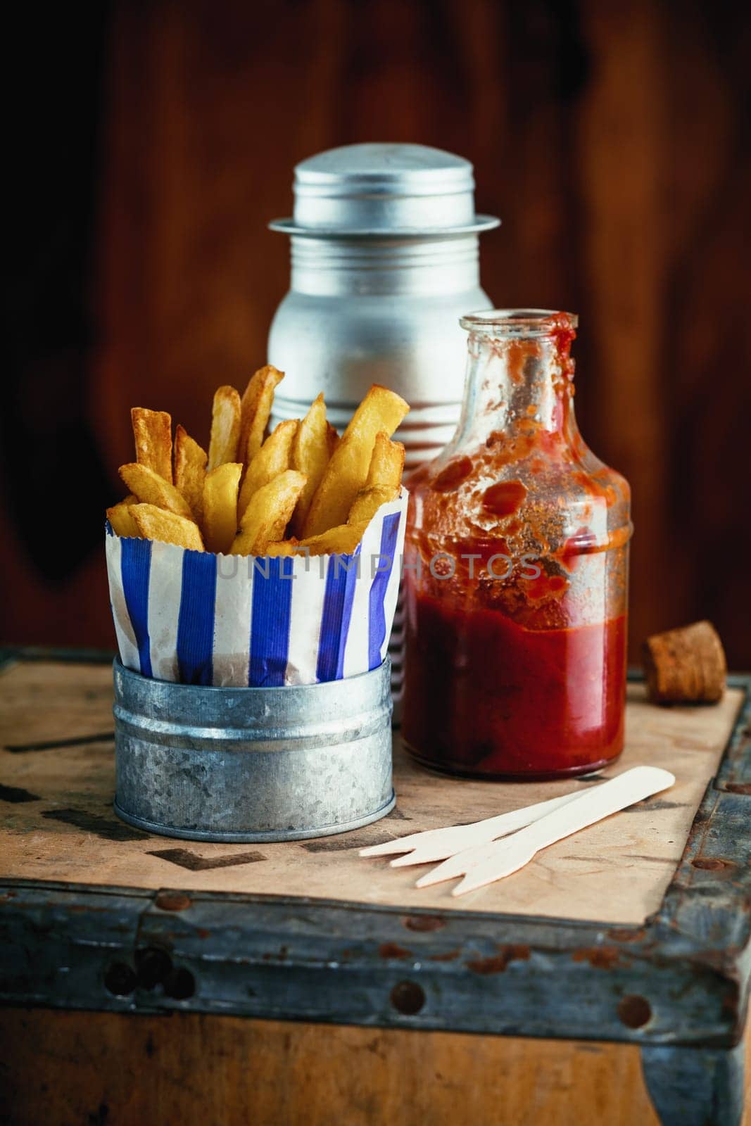 Colorful blue striped packet of crispy of French fries standing on a rustic wooden table in a fish and chip shop with condiments