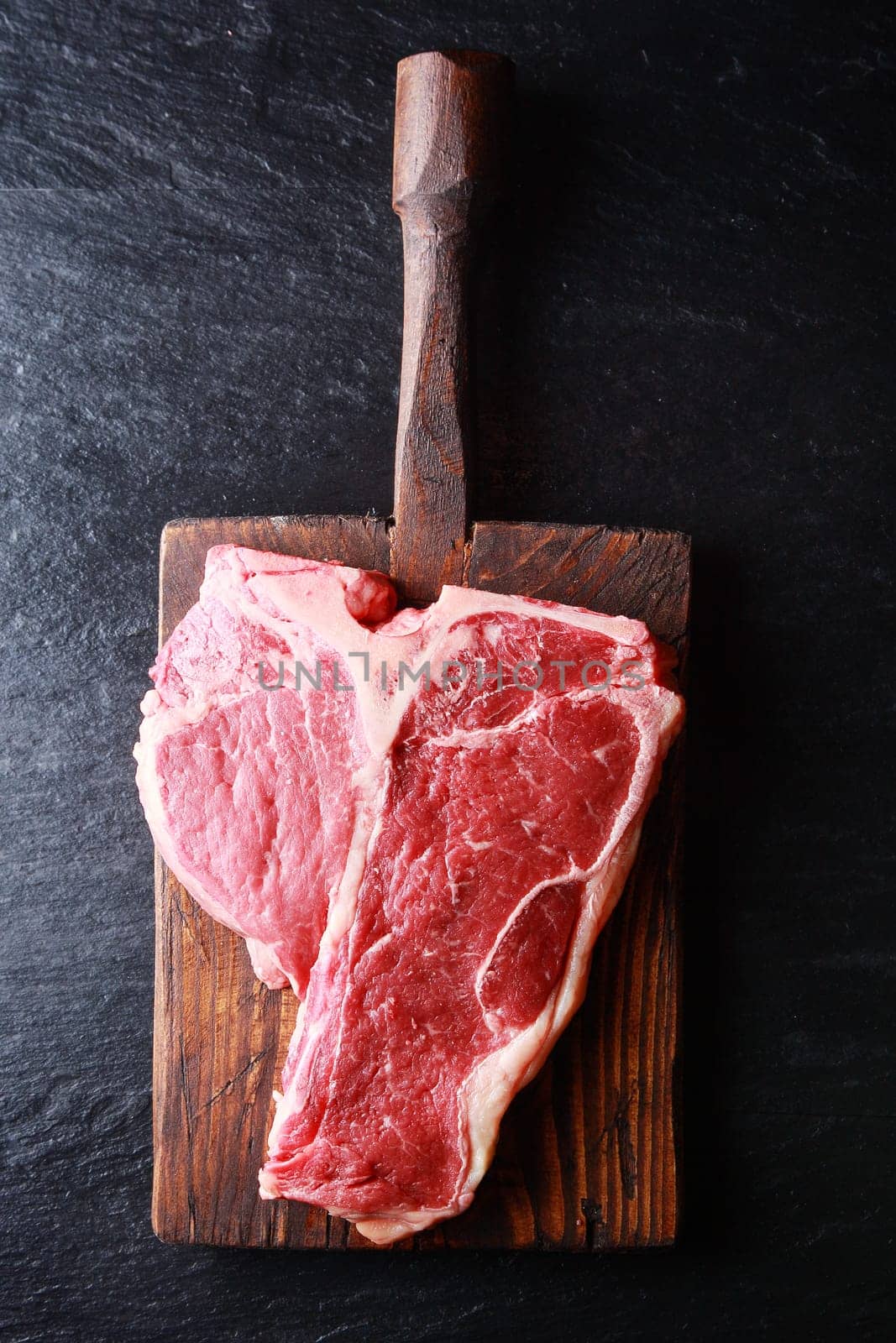 Overhead view of an uncooked t-bone steak on a rustic vintage wooden board on dark slate with copyspace