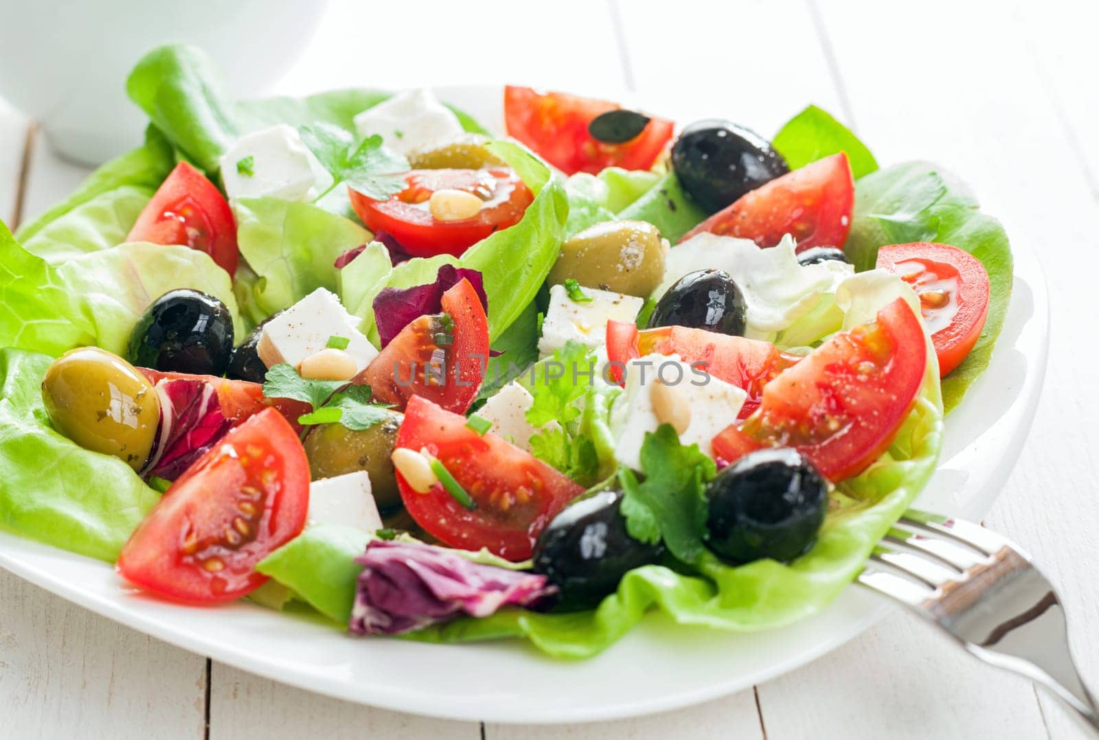 Plate of fresh salad with feta cheese, tomato , leafy greens and assorted olives on rustic white wooden boards