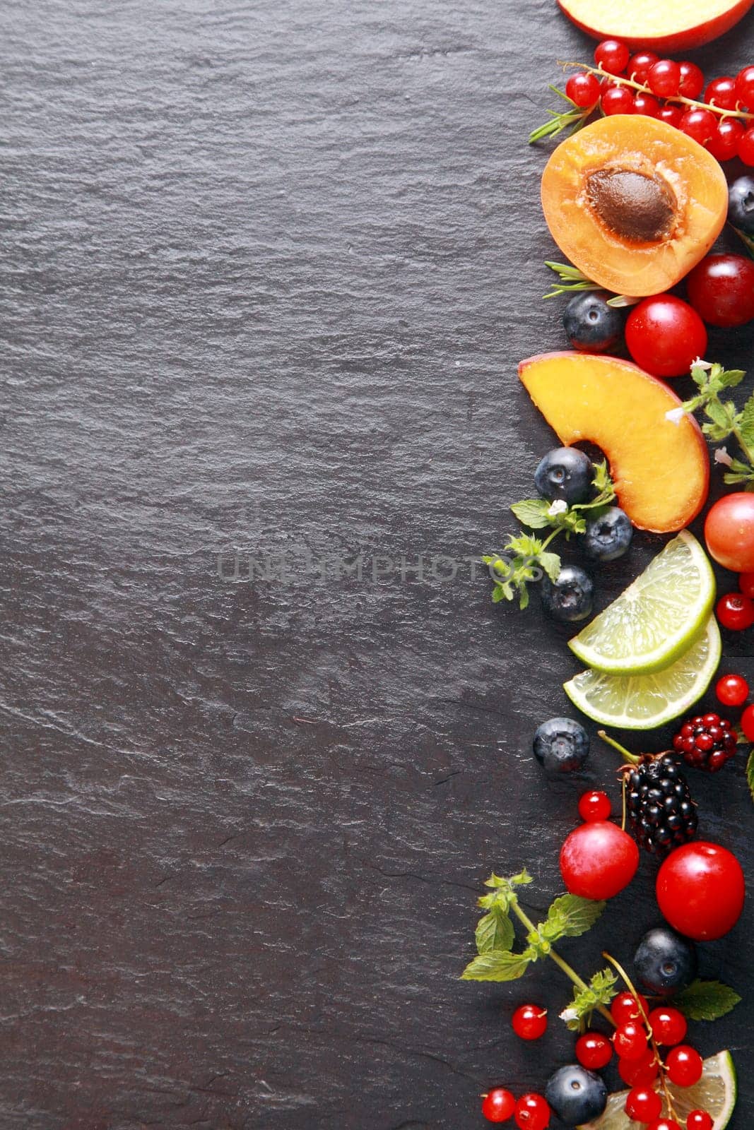 Border of fresh fruit and herbs along the right of the frame with assorted berries, apricot, peach and lemon on a dark grey slate background with copyspace