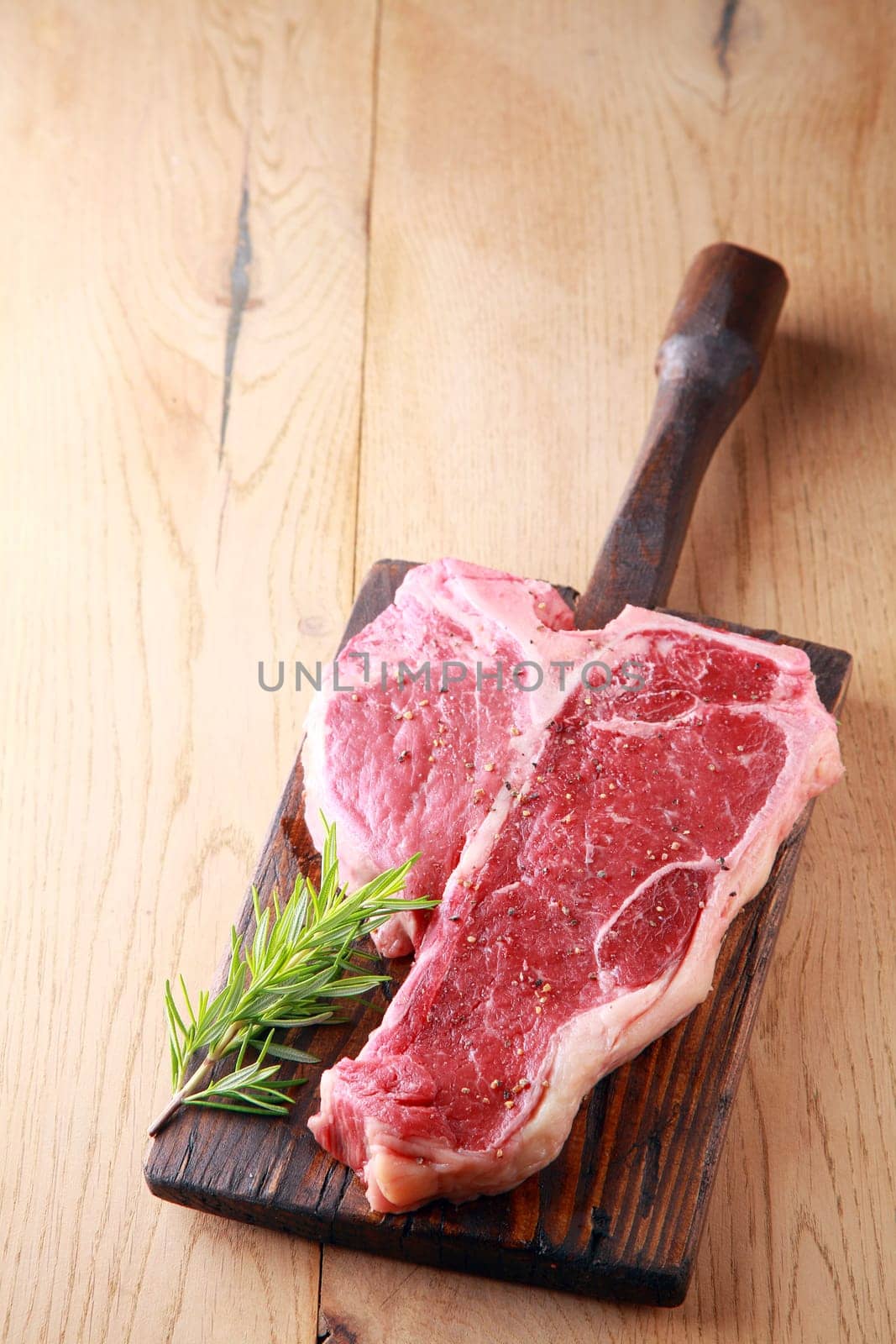 An uncooked tender lean porterhouse steak with a sprig of fresh aromatic rosemary for seasoning on an old rustic wooden board on a wood table with copyspace, view from above
