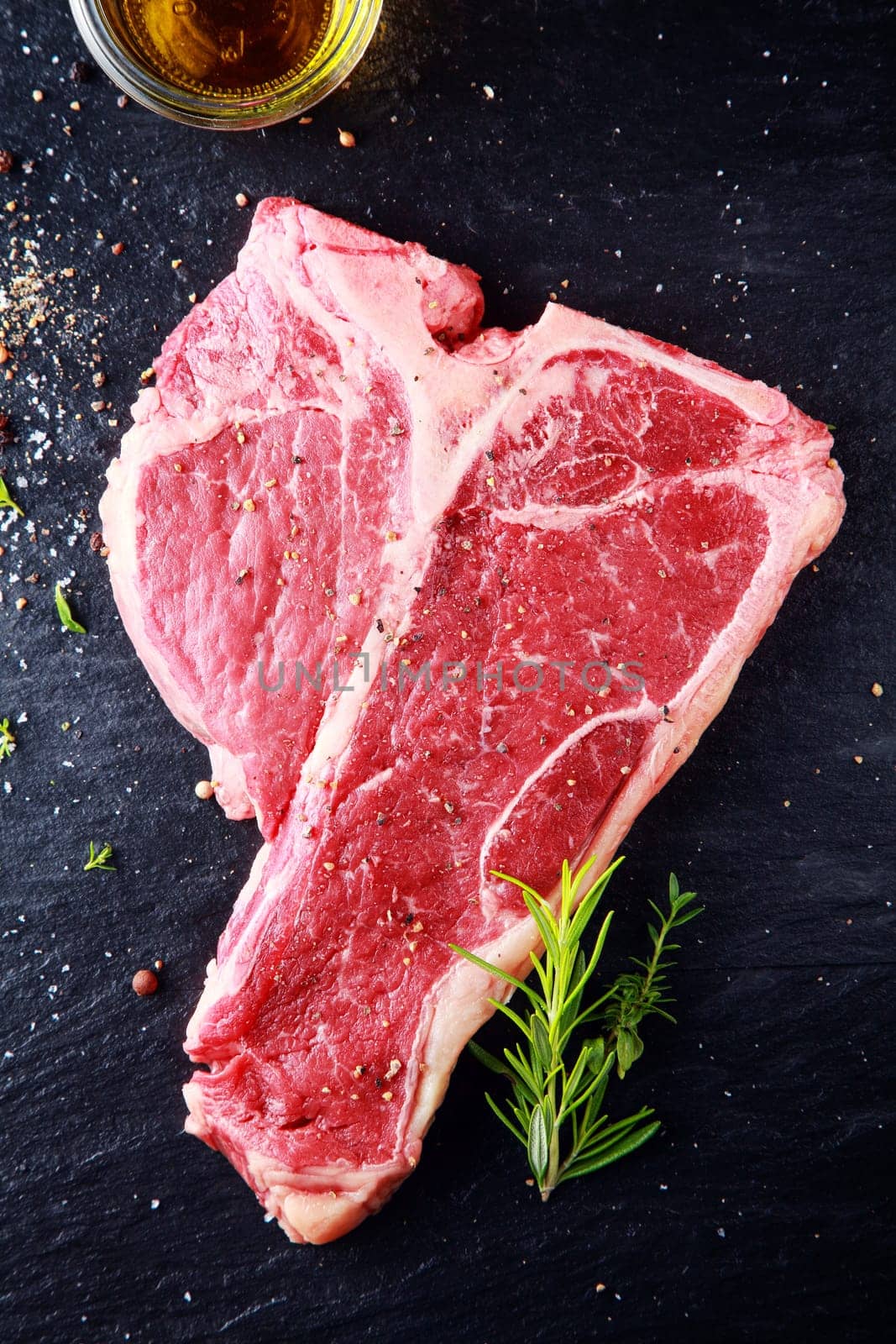 Uncooked t-bone or porterhouse steak seasoned with salt and spices with fresh herbs with a glass of whiskey above