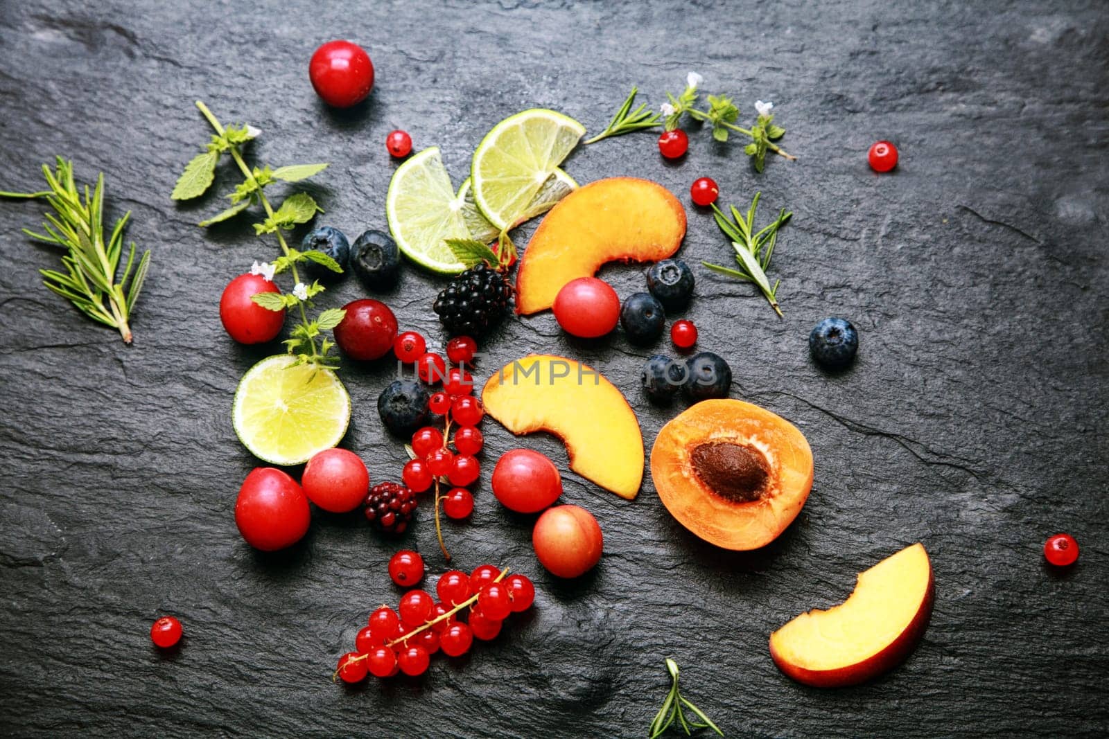 Scattered fresh fruit and berries with cranberries, blackberries, redcurrants, lemon, peach, blueberries, rosemary and herbs on a textured slate background