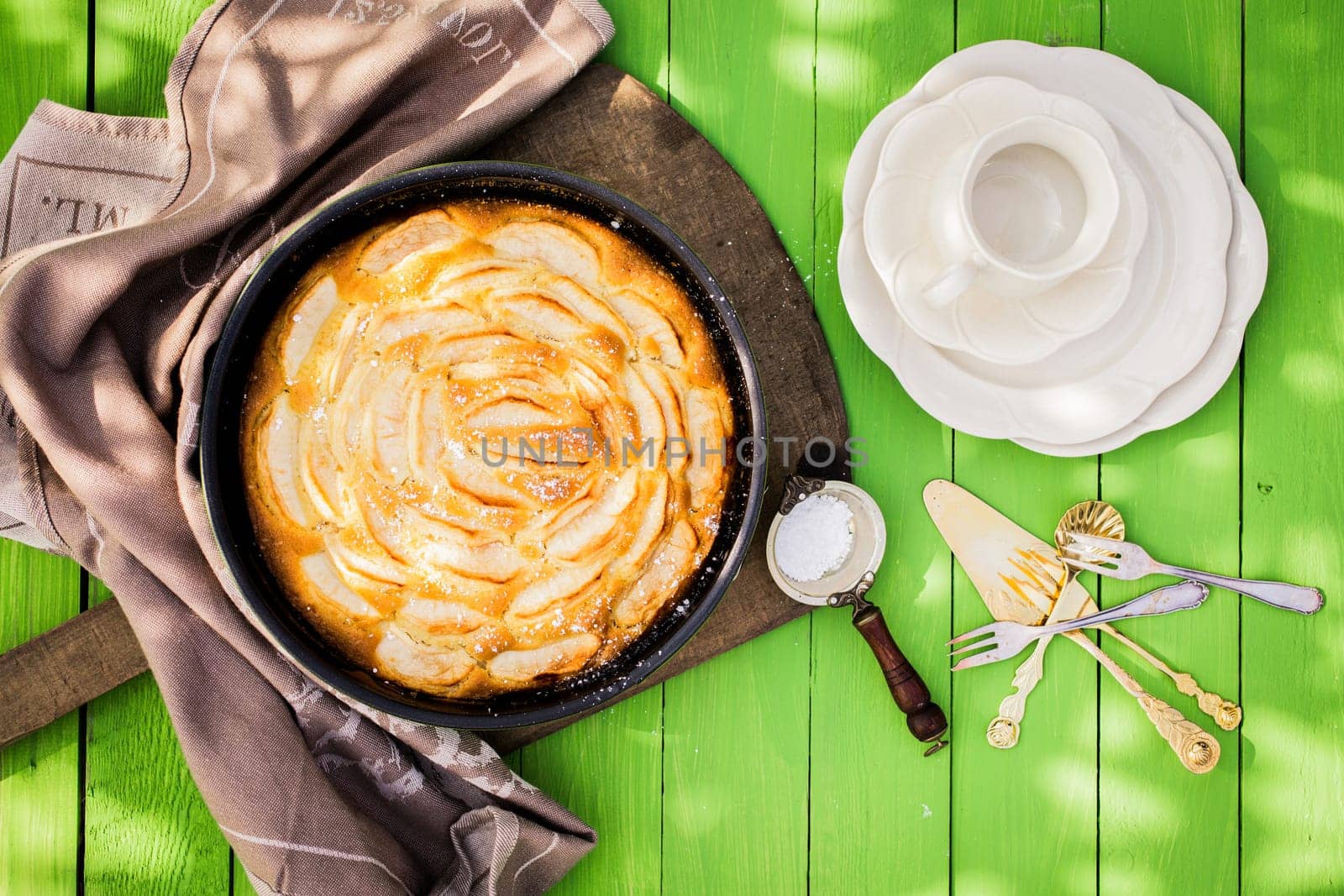 Coffee time in the garden with fresh apple pie with an overhead view of an empty cup and cake plates alongside a golden tart in a dish with a strainer of powdered sugar to sprinkle on top