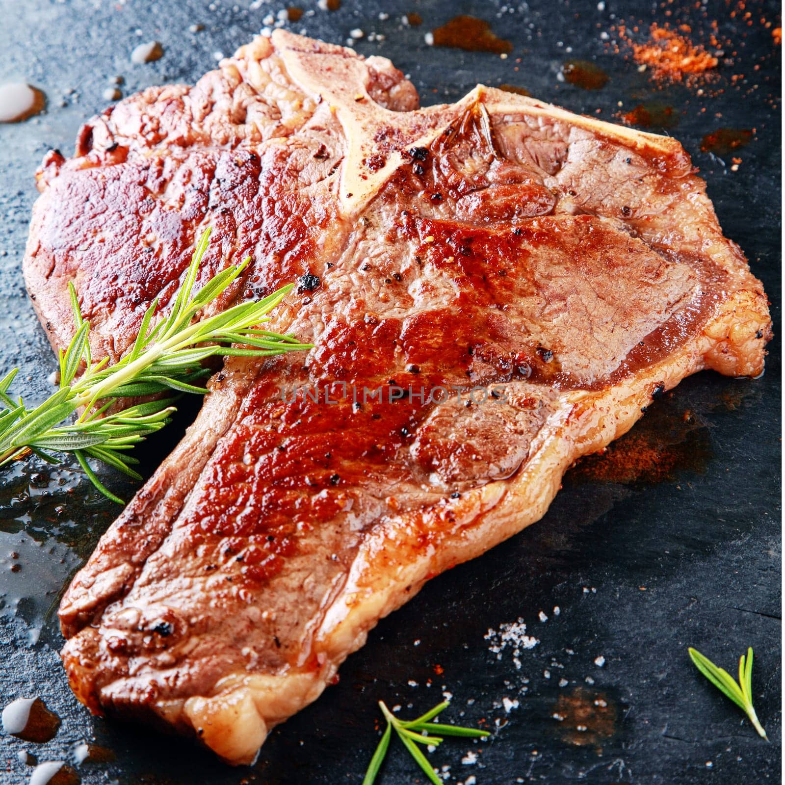 Delicious grilled t-bone steak on a hot griddle seasoned with fresh rosemary, herbs and spices in a steakhouse or restaurant