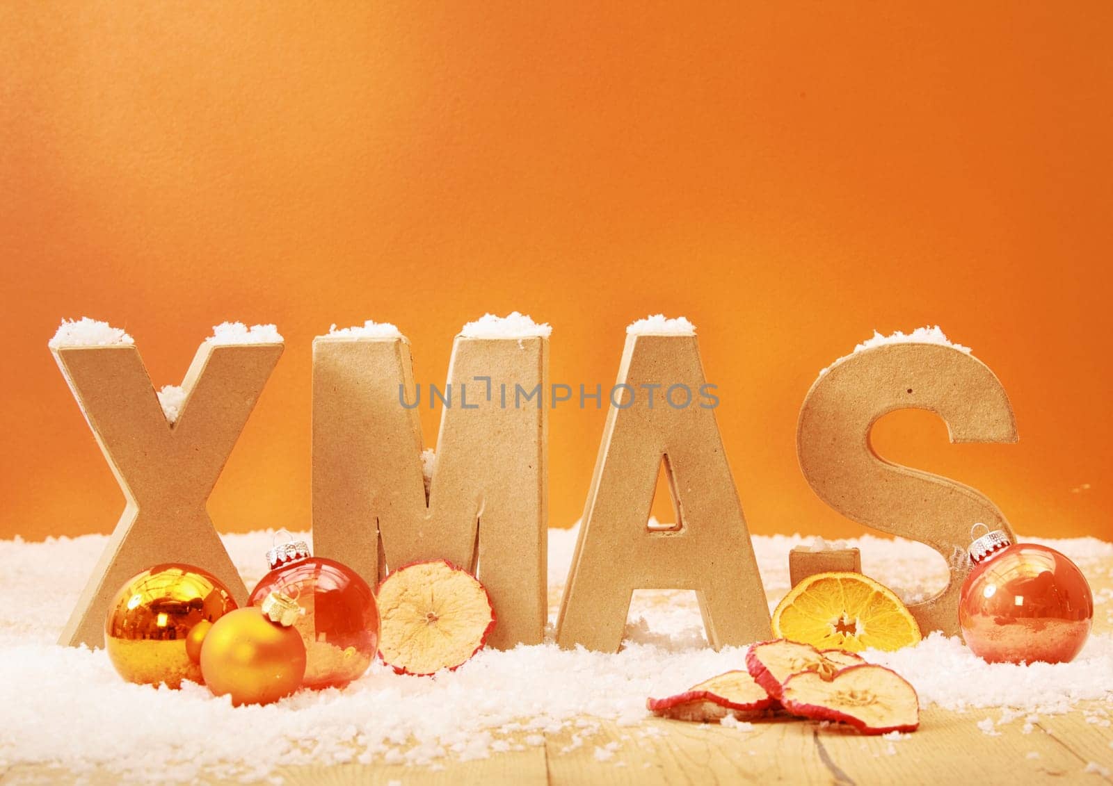 Warm toned Xmas background with wooden letters for Xmas in snow with orange and gold decorations and dried apple and orange slices against an orange background with copyspace