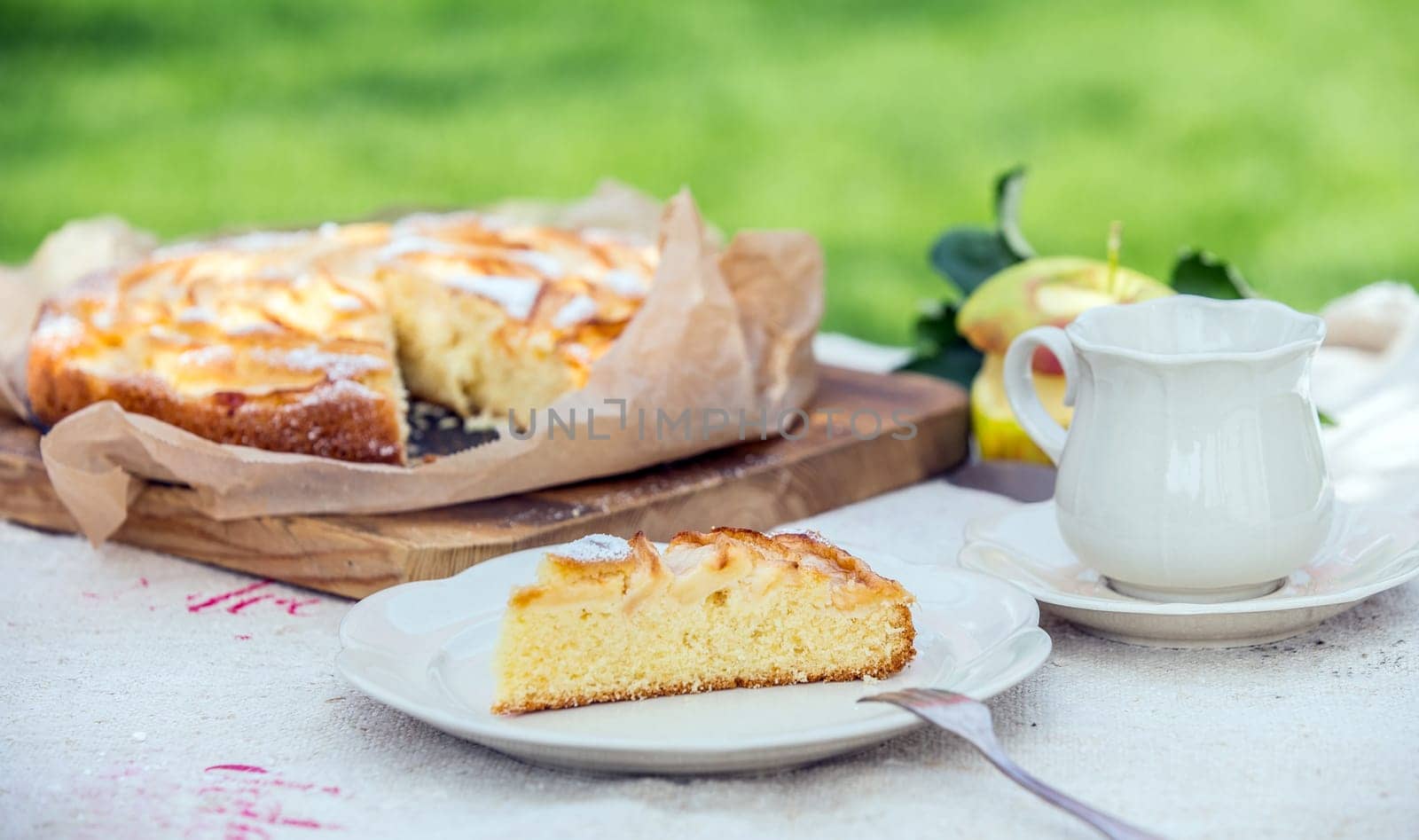 Slice of tasty freshly baked apple tart for a summer coffee or tea break in the garden with a cup and saucer and apple pie on a rustic white table