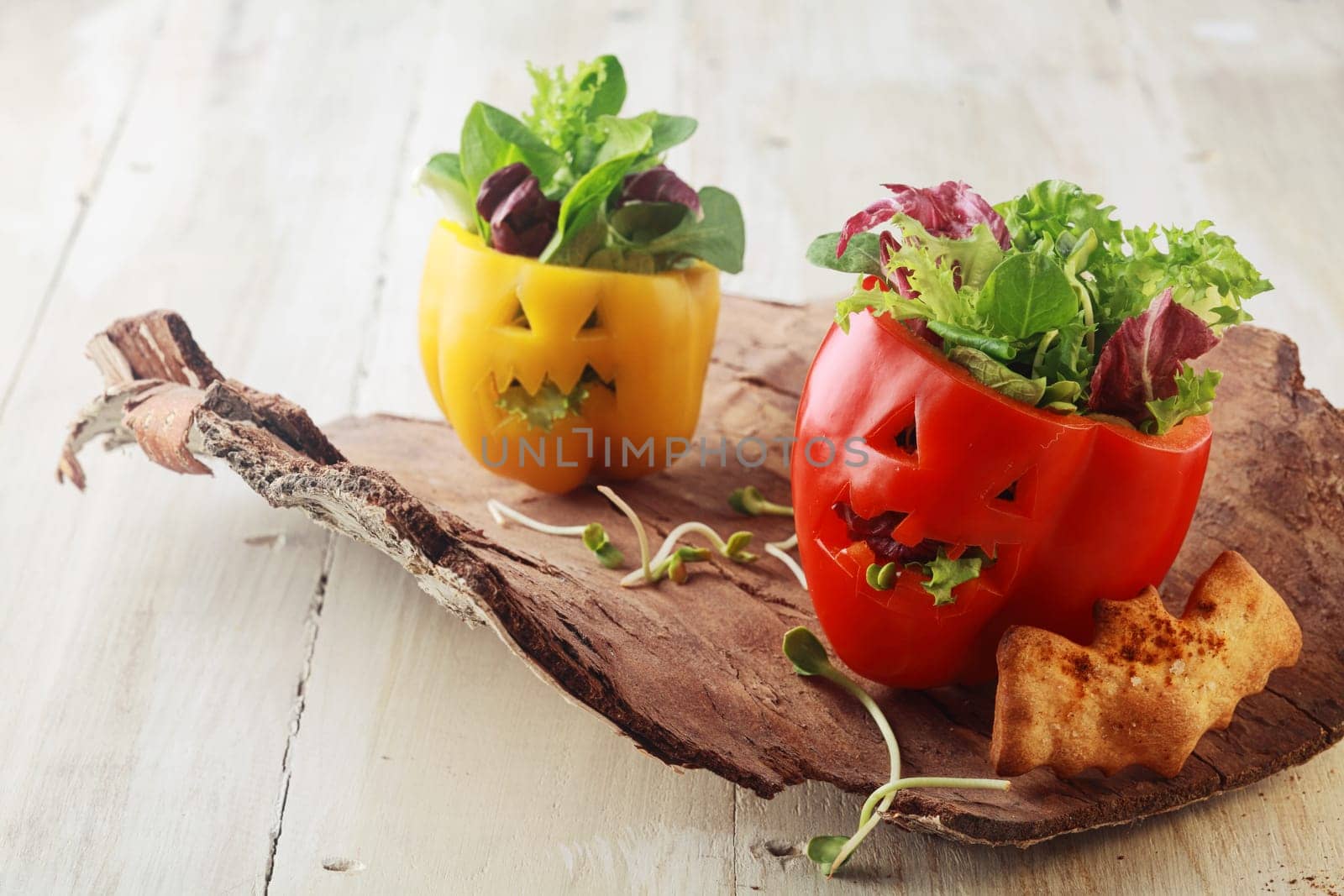 Colorful fresh Halloween bell pepper salads in individual servings for appetizers with carved jack-o-lantern faces stuffed with leafy green lettuce and herbs served on a bark platter with a bat cookie