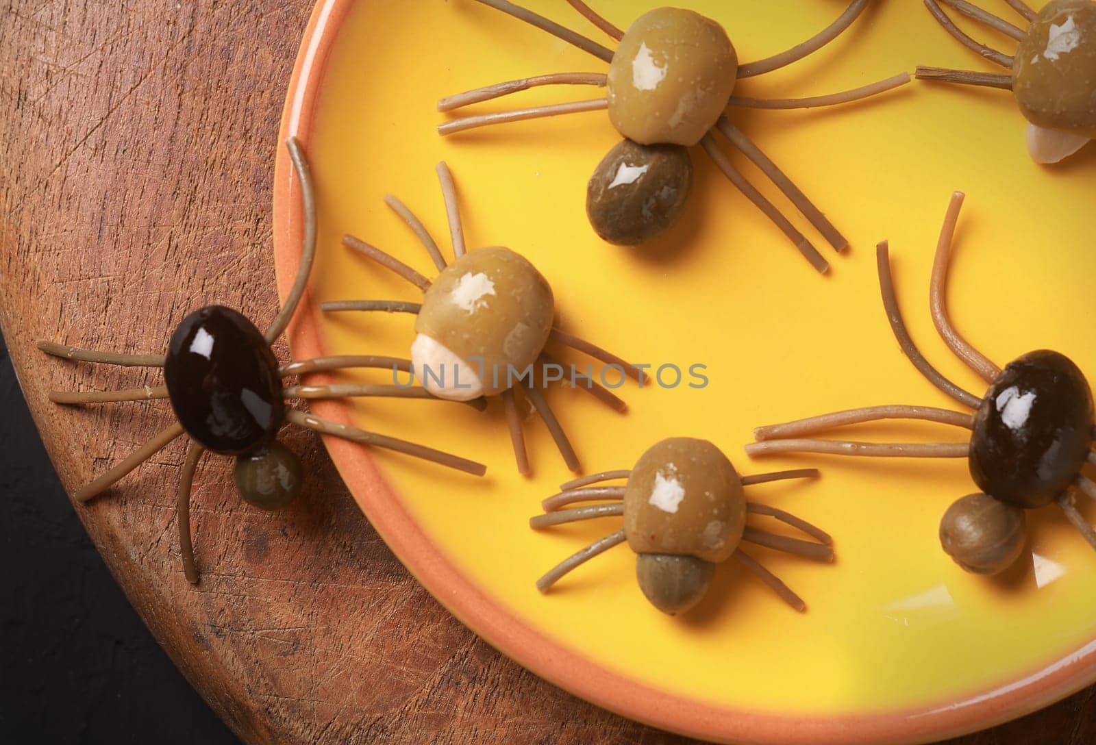 Cute spooky Halloween spider appetizers made from green, black and stuffed olives with spaghetti legs crawling off a yellow platter onto a wooden table, view from above