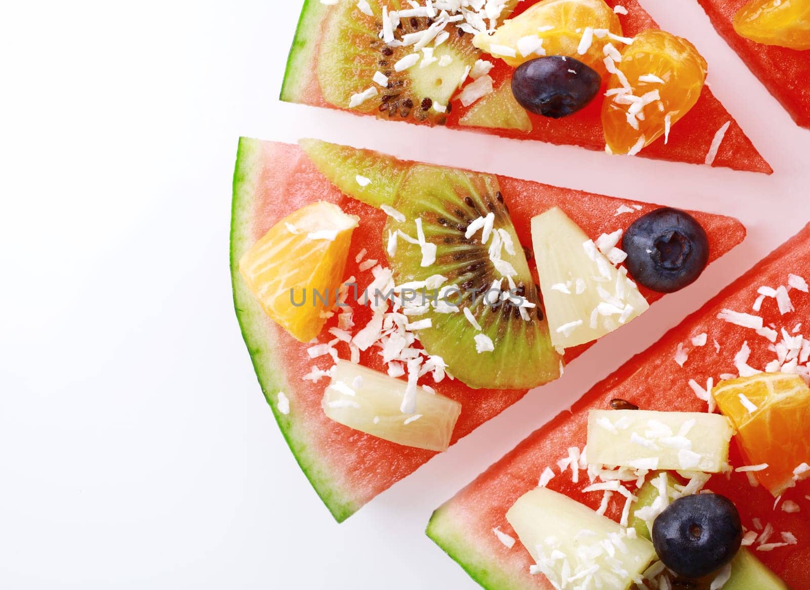 Close up overhead detail of a slice of tropical fruit watermelon pizza topped with kiwifruit, blueberries, orange, pineapple, and sprinkled with desiccated coconut, on white with copyspace
