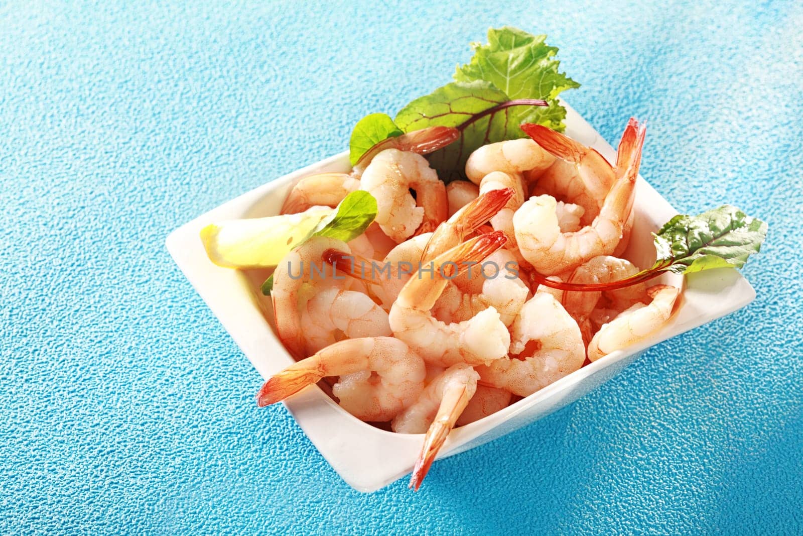 Gourmet pink prawn or shrimp tails served in a dish with lemon and fresh herbs for a delicious seafood appetizer to a dinner, high angle view on turquoise blue with copyspace