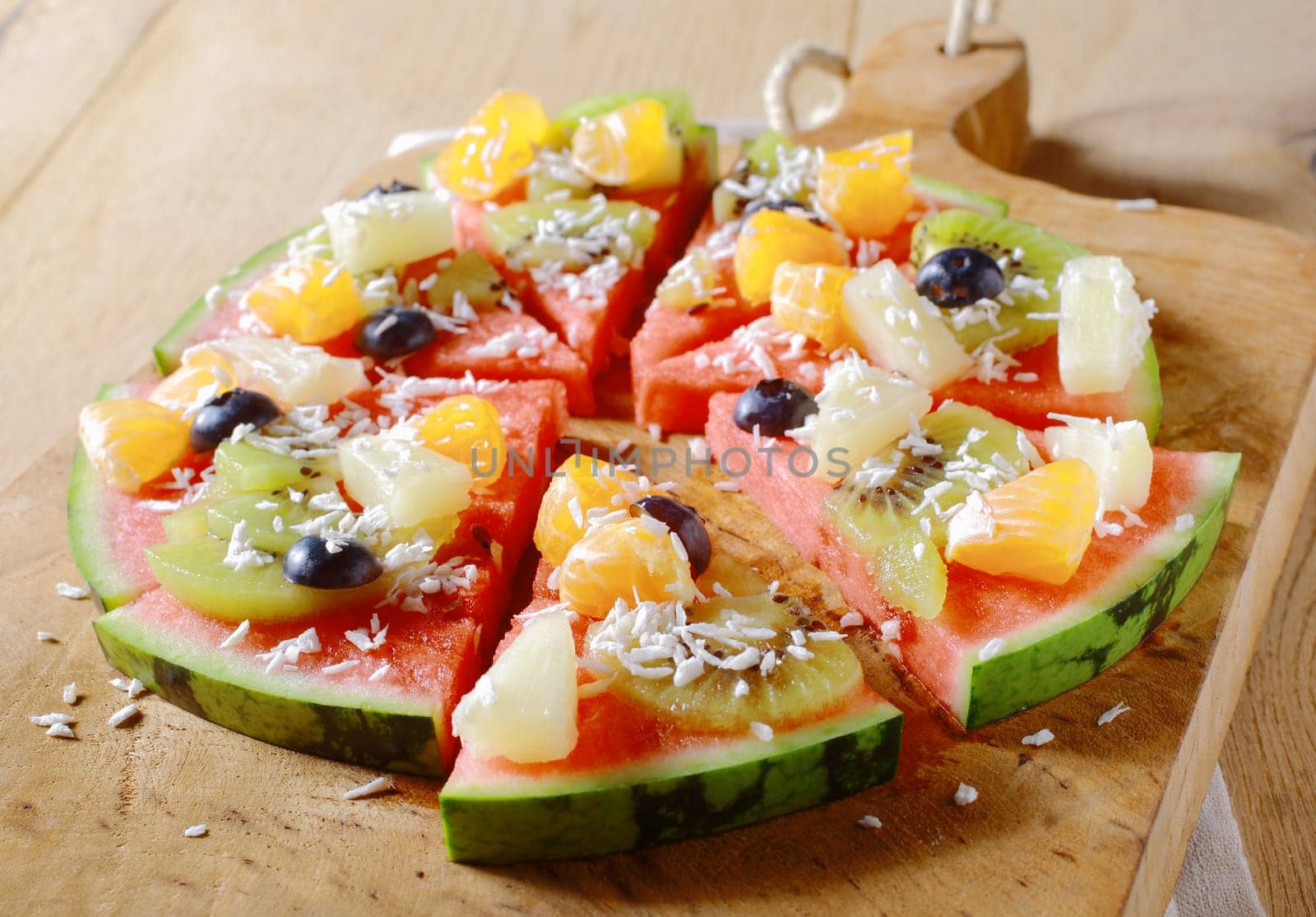 A tasty juicy fresh tropical fruit watermelon pizza topped with kiwifruit, blueberries, orange, pineapple, and topped with dried coconut, cut into segments on a wooden board for a nutritious dessert