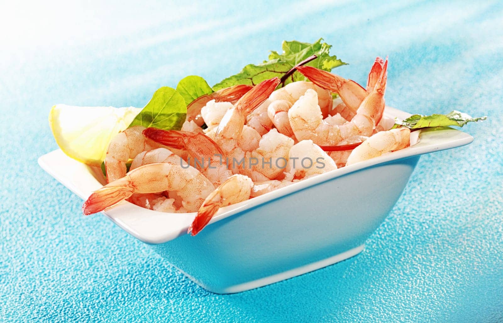 Delicious seafood appetizer of grilled shrimp or pink prawns served in a dish with a slice of lemon and salad greens on a turquoise blue background