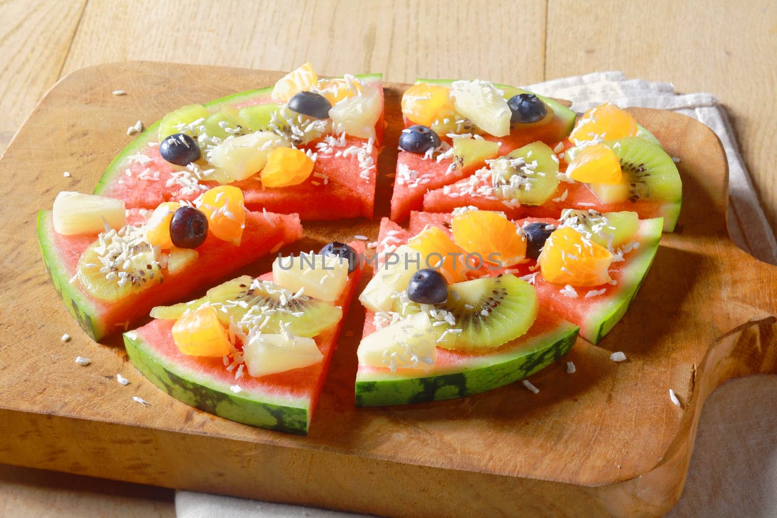 Low angle view of a healthy vegetarian and vegan tropical fruit watermelon pizza topped with kiwifruit, blueberries, orange, pineapple, and sprinkled with dried coconut, cut into segments for serving