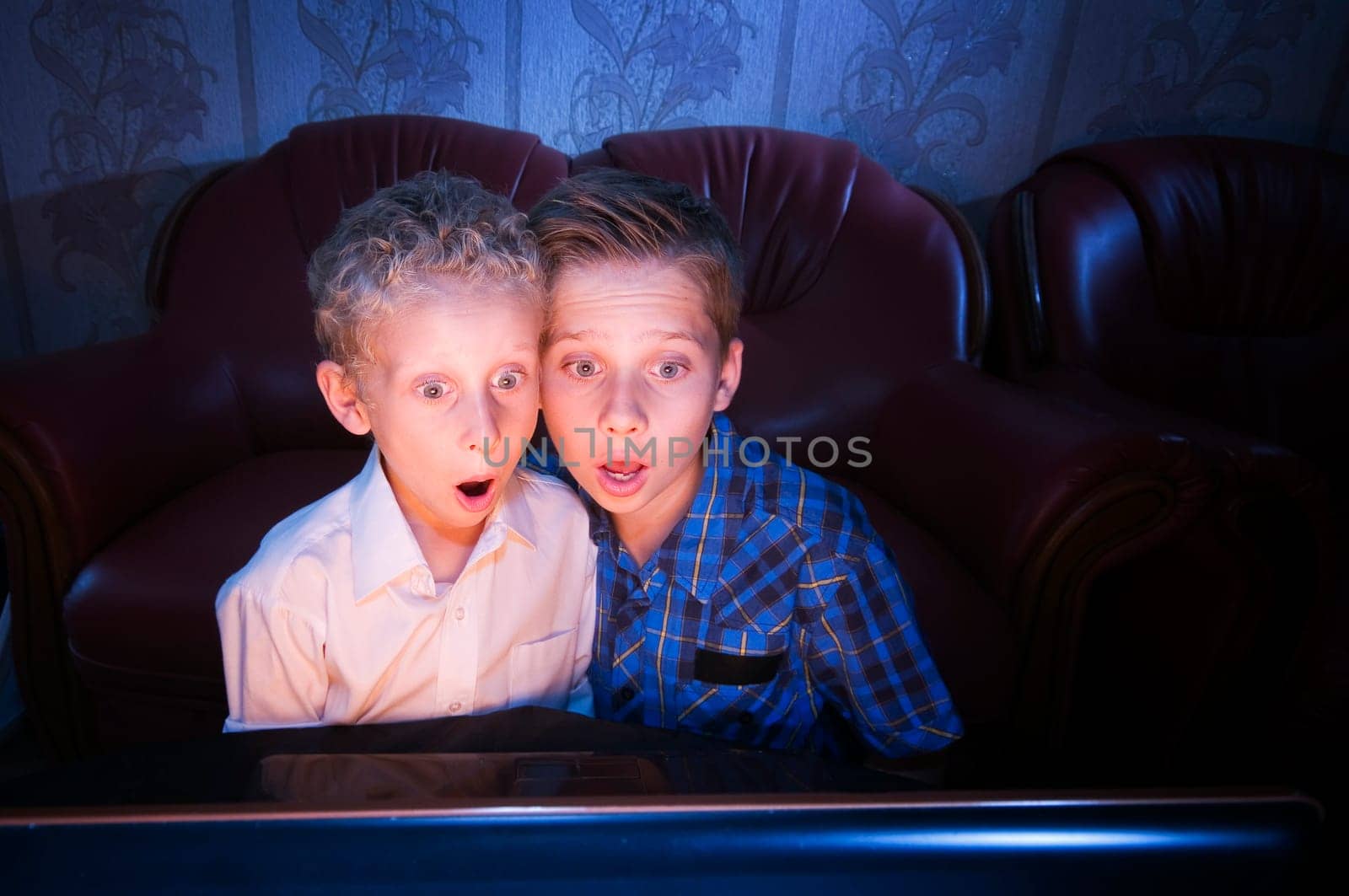 Frontal view of two brothers aged 7 and 13 in the darkened room shocked looking at the TV