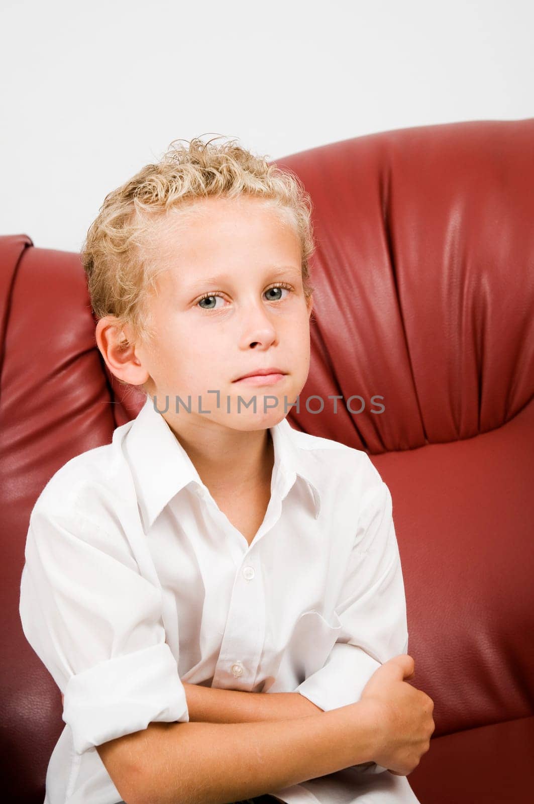 Upper body portrait one seven year old blonde curly boy on brown leather sofa against white background
