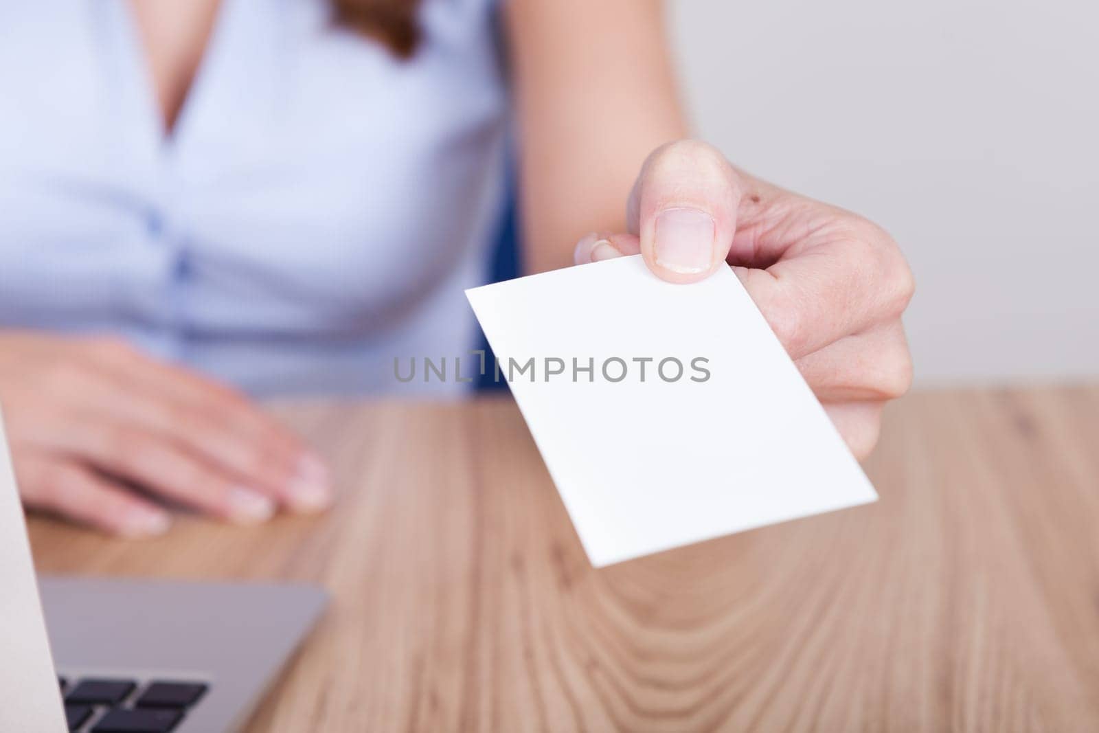 Young businesswoman sitting behind her laptop holds out her hand offering her card with focus to her face