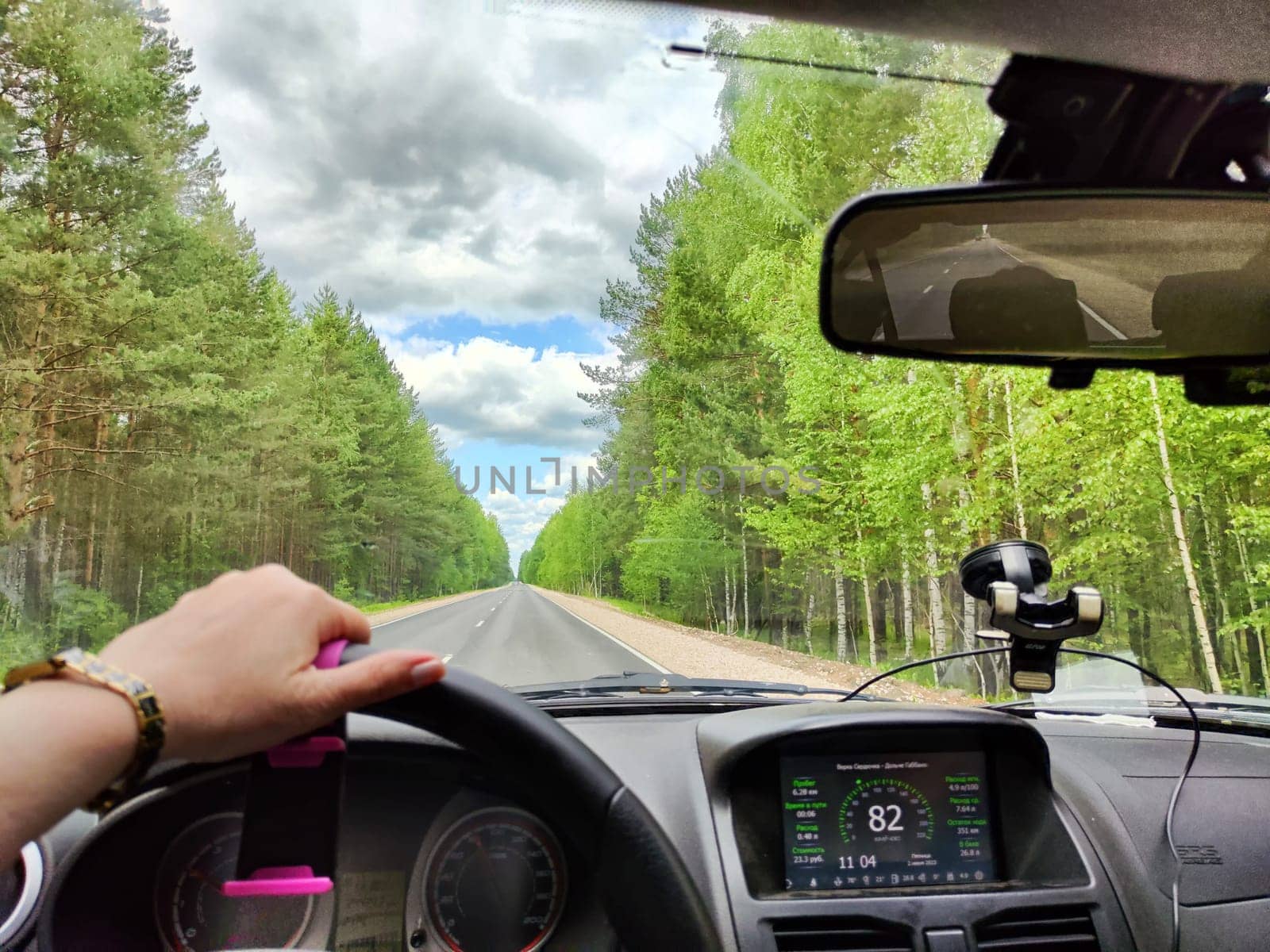 view through car windshield of natural landscape with road, green tree, white clouds. Hand of woman on steering wheel of car. Female traveler driving on trip or journey in spring or summer