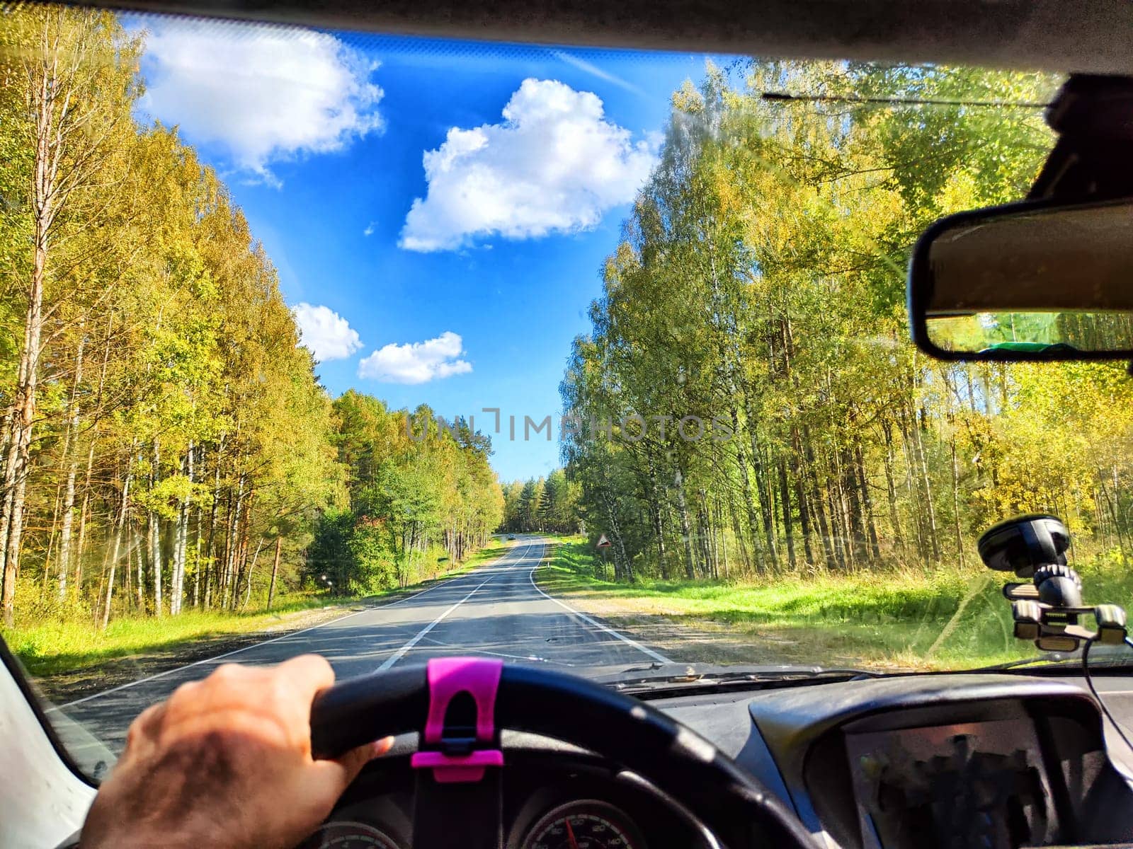 view from a car windshield of natural landscape with road, green trees and blue sky in summer or spring time. Hand of woman on steering wheel. Female traveler driving alone on trip or journey