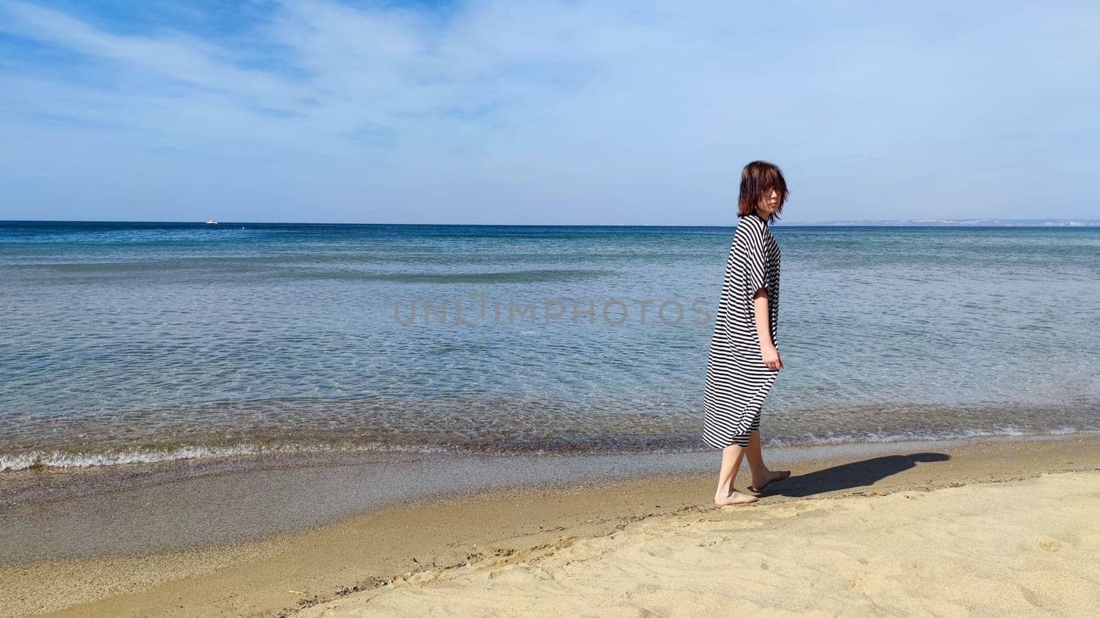 Teenage girl in a striped dress on the seashore on a sunny day by Annado