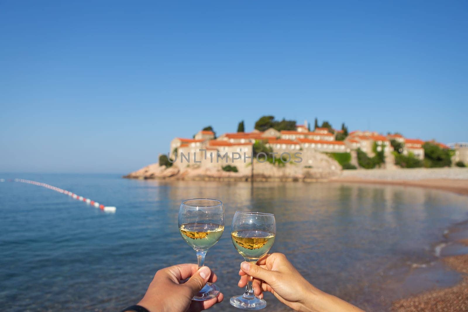 Sveti Stefan Island, Montenegro July 5, 2021: Adriatic Sea. Glasses with wine on the background of the island. Romantic trip. by sfinks