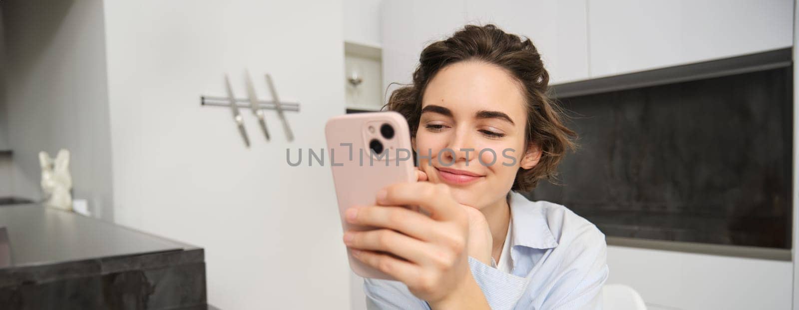 Portrait of woman sitting at home and checking her mobile phone, looking at smartphone with happy smile by Benzoix