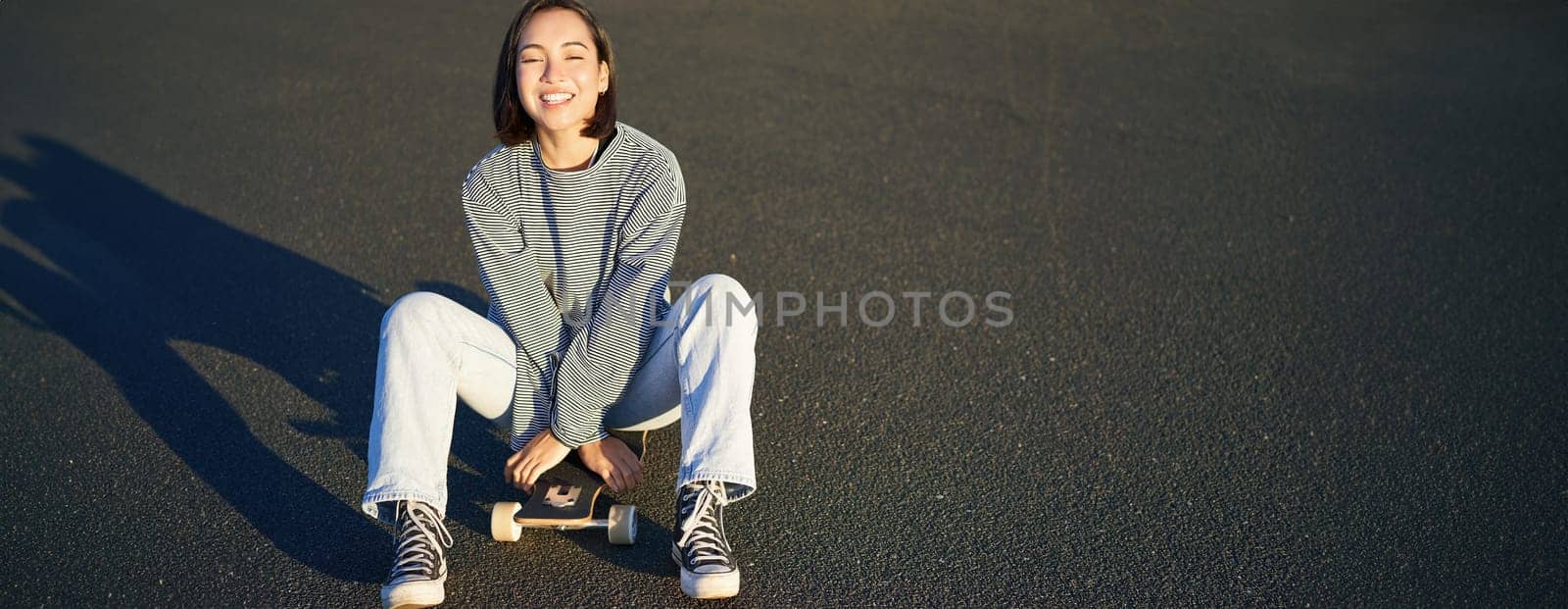 Freedom and happiness. Cute smiling asian girl, sits on skateboard on sunny spring day. Happy laughing skater enjoying cruising on longboard by Benzoix
