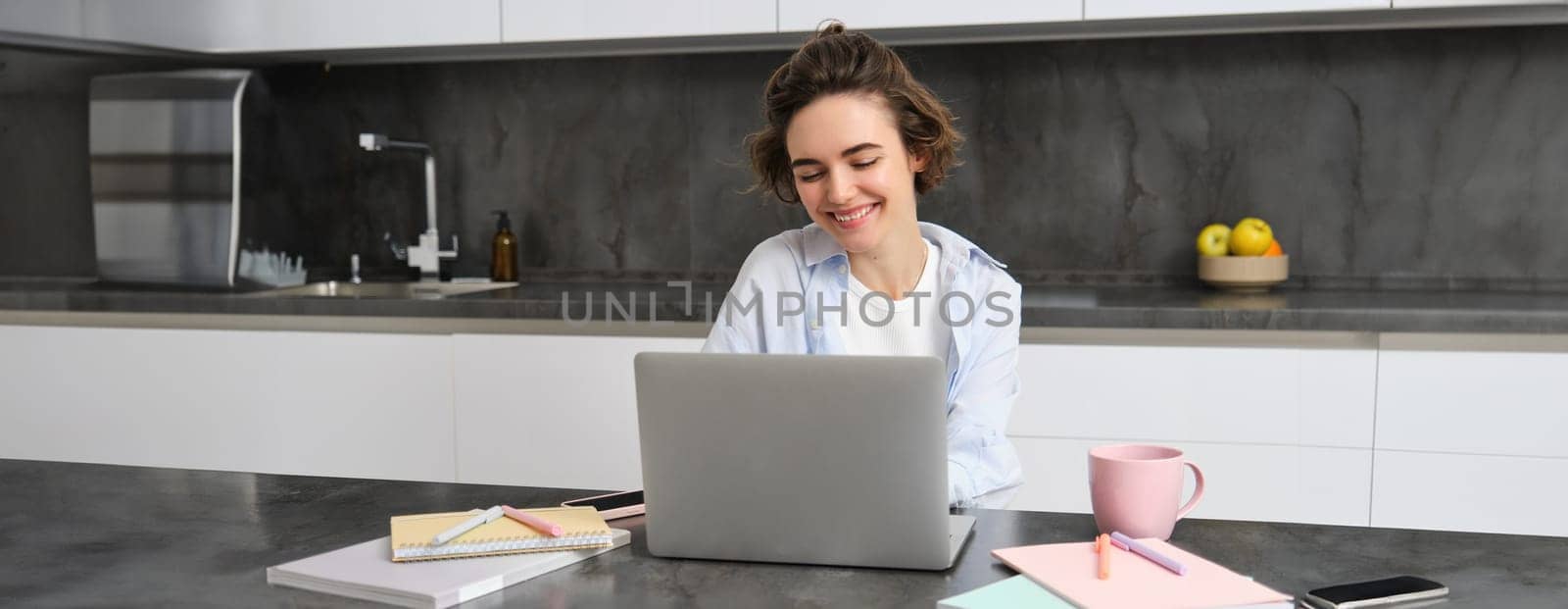 Image of young productive woman works from home, does distance learning, learns online course on her laptop in kitchen, writes down information, watches webinar, attends work meeting remotely by Benzoix