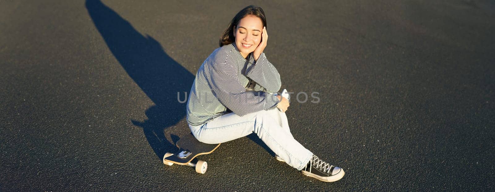 Portrait of asian woman sitting on skateboard, skating on her cruiser longboard, using smartphone app by Benzoix