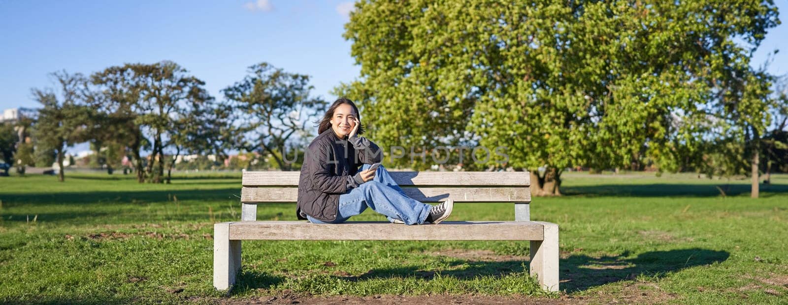 Portrait of young asian woman sitting on bench with smartphone. Cute girl using mobile phone, enjoying being outdoors.