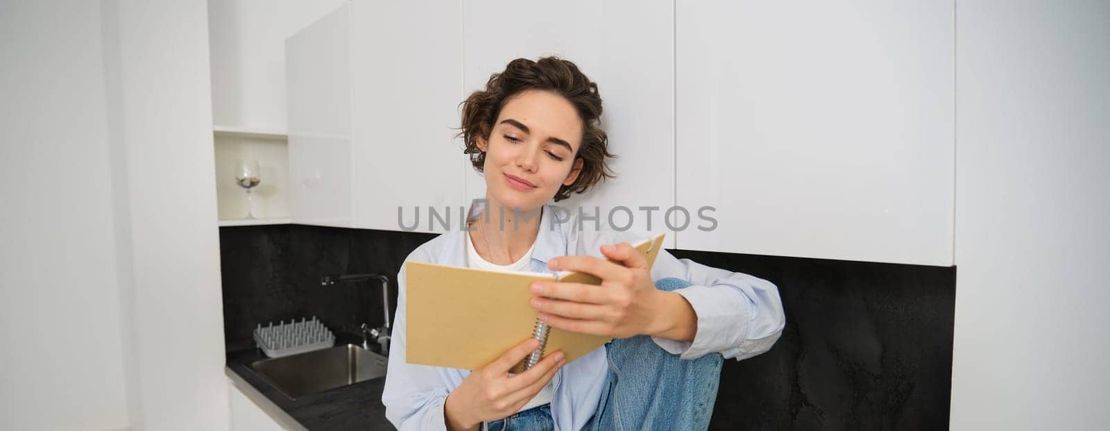 Portrait of smiling young woman reading journal, enjoys comfort at home, holding notebook, looking happy, relaxing indoors.