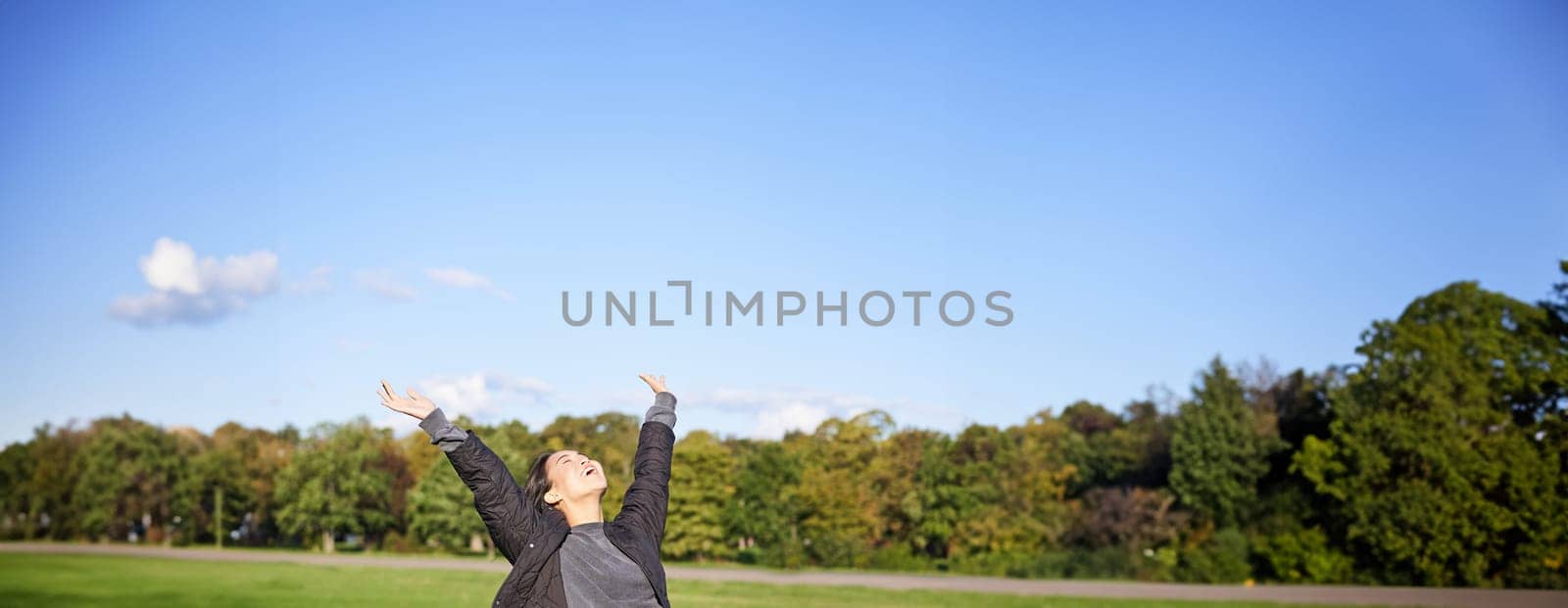 Happy asian woman stretching her hands, sitting on bench with excited face, smiling pleased, feeling freedom, enjoying day in park.