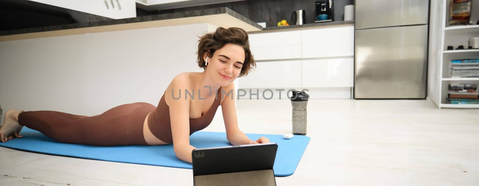 Young fitness woman, girl in tracksuit lying on fitness mat, looking at yoga, pilates video tutorial on tablet, does workout from home, uses gym app, training at home.