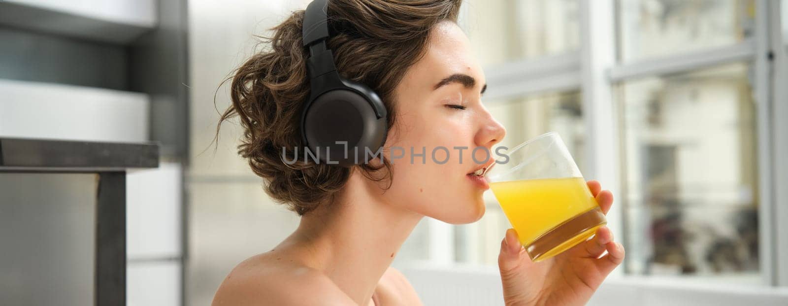 Smiling sportswoman, fitness girl does workout at home, wears headphones, listens music and drinks orange juice after training session.