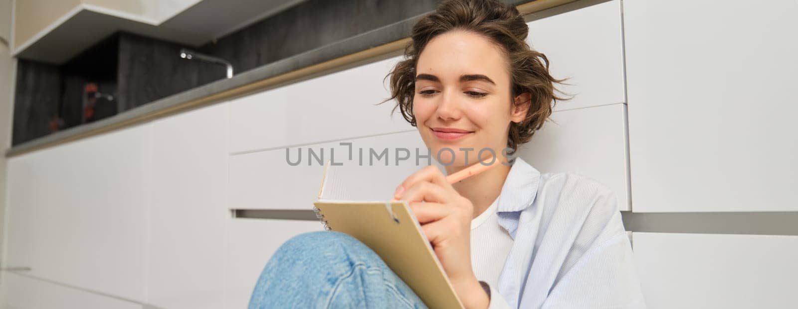 Hobbies and lifestyle concept. Happy young woman sits on floor with pen and notebook, wirtes in her diary, makes notes in her journal, plans her schedule, draws on paper by Benzoix