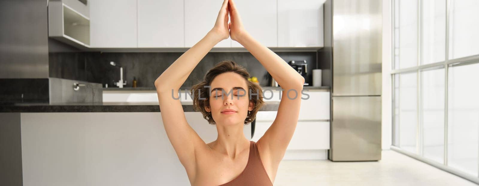 Image of young woman meditating, raising her hands up above head, practice mindfulness at home, doing yoga session on rubber mat in bright room, wearing sportswear.