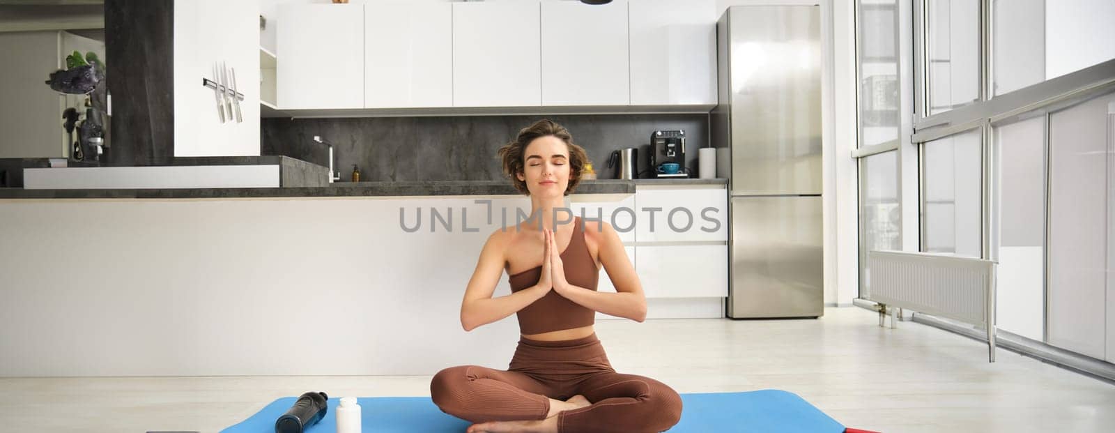 Smiling girl athlete, sportswoman doing yoga at home in activewear, sitting on yoga rubber mat in lotus pose, meditating, practice mindfulness exercises by Benzoix