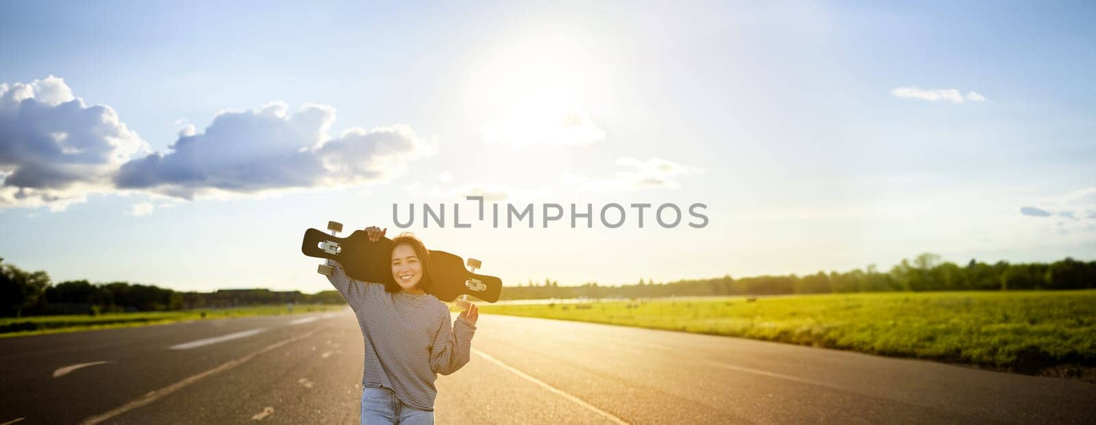 Young asian woman standing with longboard on sunny road, skating in skate park on her cruiser. Copy space