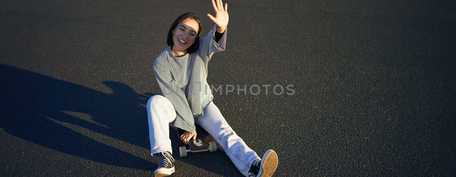 Positive korean girl covers her face from sunlight, sits on skateboard and smiles happily by Benzoix