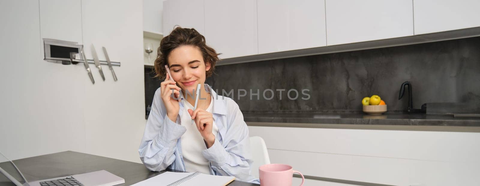 Image of woman working at home, making a phone call, sitting with smartphone, surrounded with paperwork, doing homework and talking to someone.