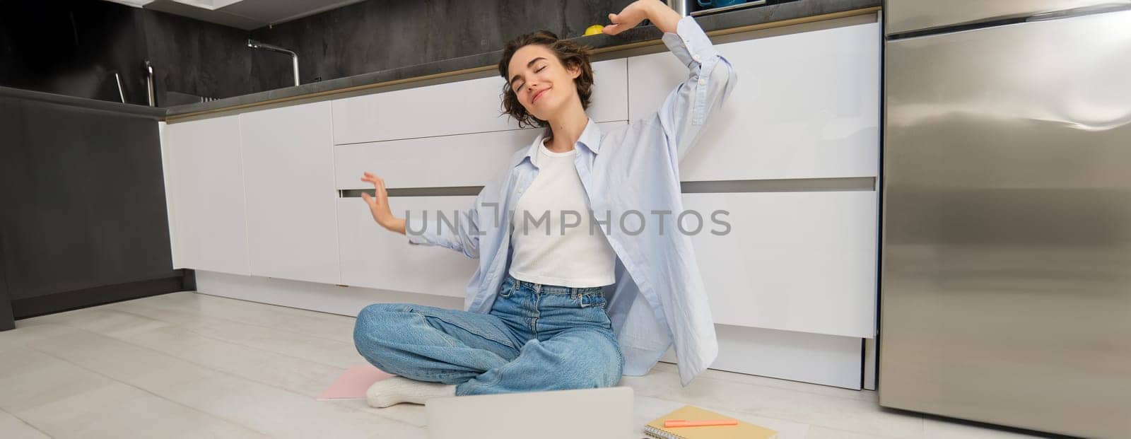 Happy young woman stretches her arms, sits on floor with laptop, finishes her remote work, completes homework on computer by Benzoix
