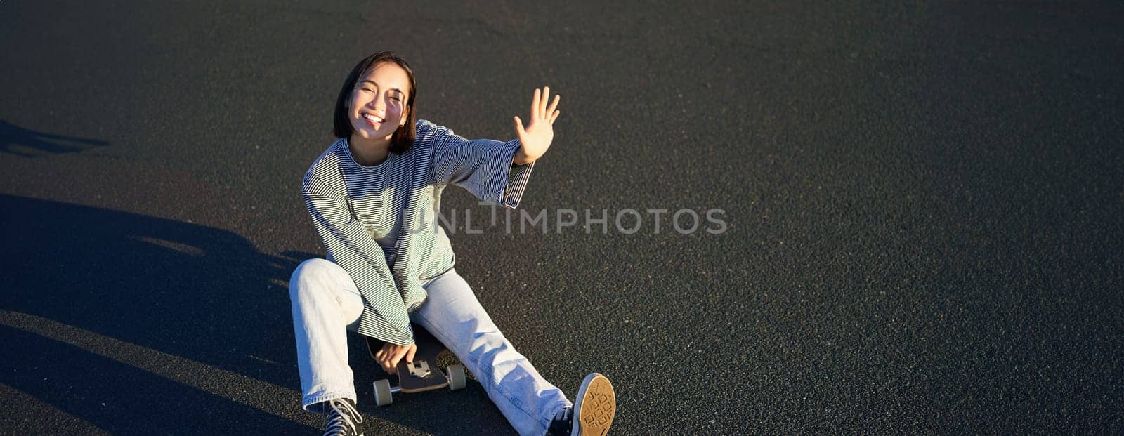 Positive korean girl covers her face from sunlight, sits on skateboard and smiles happily by Benzoix