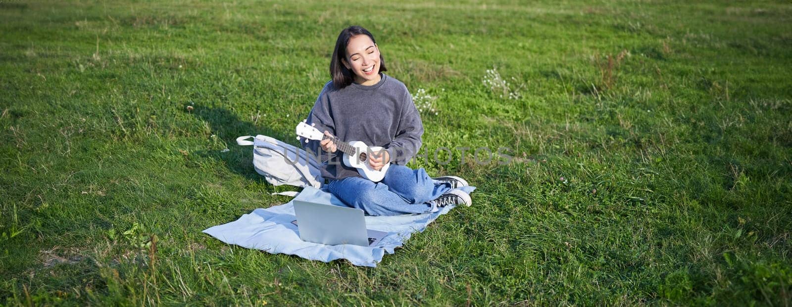 Cute korean girl, musician sits in park, plays ukulele and sings, looks up chords and tutorials on laptop.