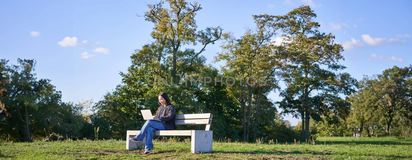 Portrait of young student, girl using laptop, sitting in park on bench, typing on computer.