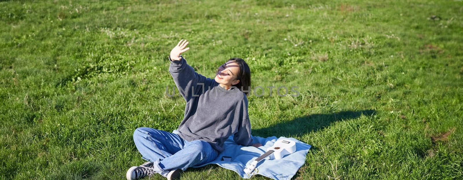 Happy asian girl sitting on picnic blanket with ukulele, cover herself from sunlight, extending hand towards sun beams and smiling by Benzoix