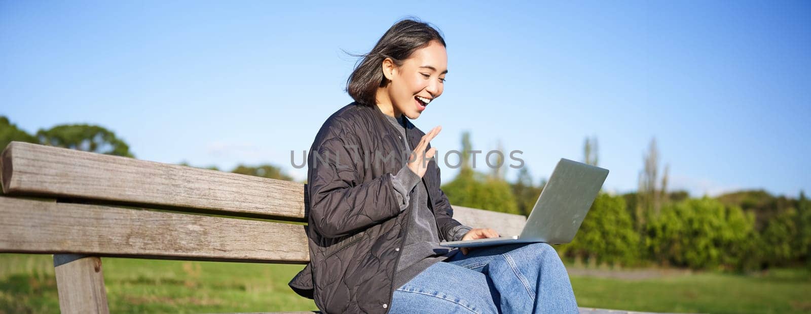 Portrait of young asian woman working remotely from park, sitting on bench with laptop, talking during video chat by Benzoix