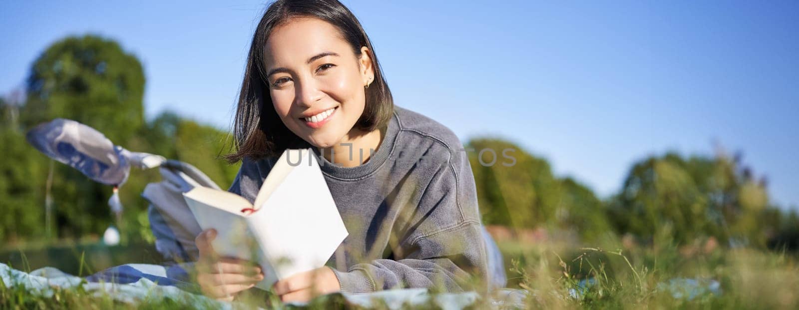 Portrait of cute korean girl, reading in park while lying on grass, relaxing with favorite book in hands, smiling happily by Benzoix